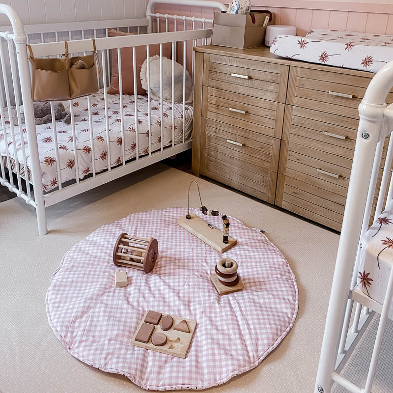 A side view of a Baby nursery featuring the Snuggly Jacks Summer Storm Baby cot sheet and Change Mat cover in a modern baby bedroom. There is a Peachy Pink Gingham Playmat on the ground with scattered wooden toys.