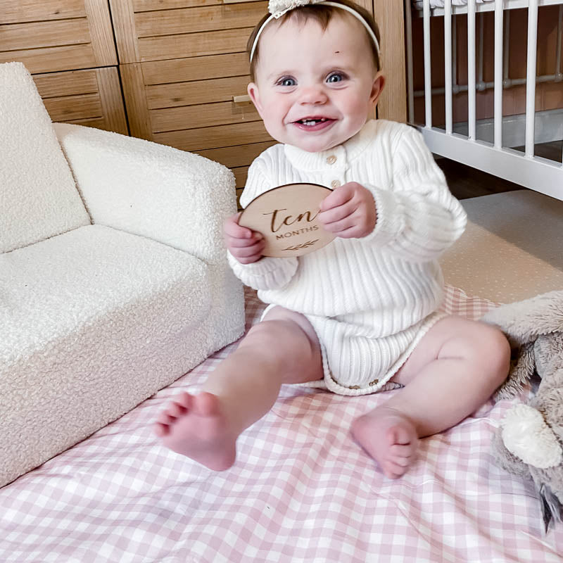 Adorable 10 month old baby girl holding a milestone disc sitting on a cotton playmat in the peachy pink gingham print from Snuggly Jacks Australia