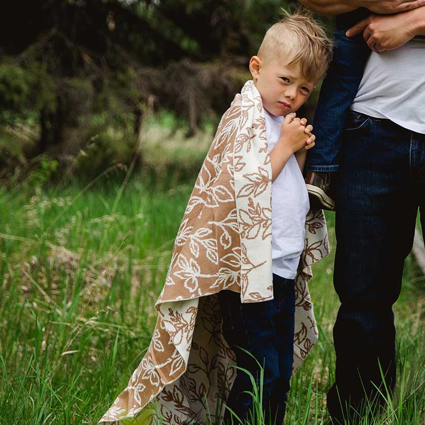 Small boy standing in a grassed paddock with a Foliage Sand Organic Knitted Blanket draped over him