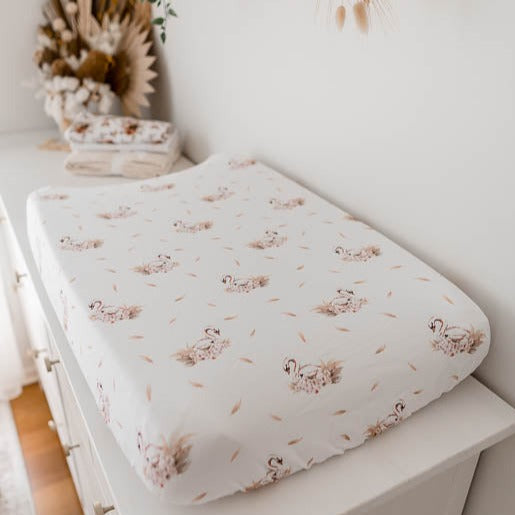 White change pad cover with soft pink swans placed in a seamless pattern set out on a whit pine change table.